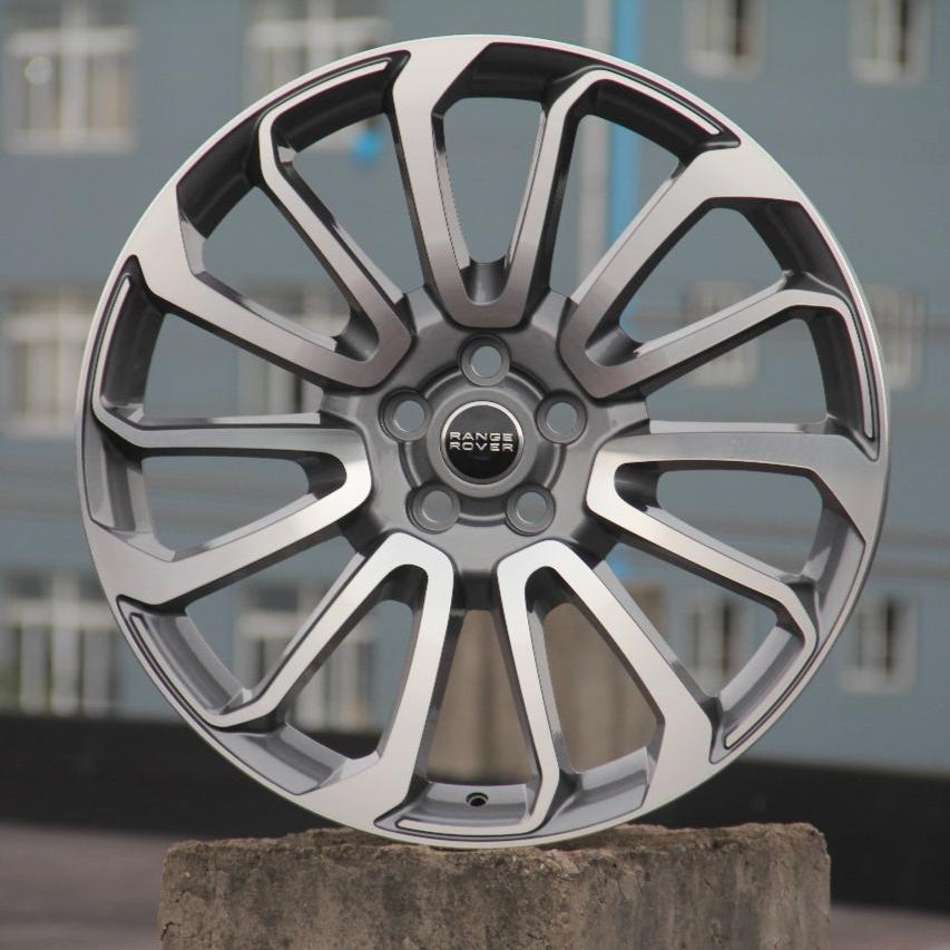 A silver alloy wheel with a multi-spoke design is displayed. The center features the "RANGE ROVER" logo. It's resting on a concrete surface against a blue building with windows.