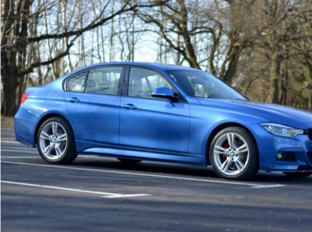 A blue car is parked in an outdoor parking lot. The scene is set in a wooded area with bare trees, suggesting early spring or late autumn.