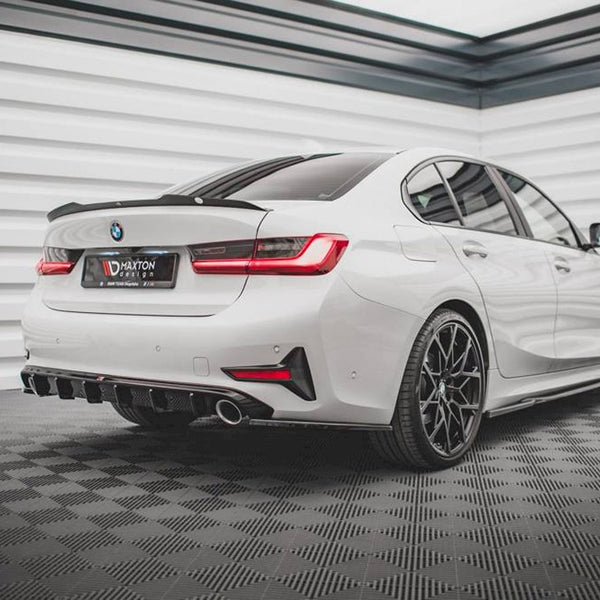 A white BMW sedan stands in a modern garage with a checkered floor. The rear features black diffuser accents and dual exhaust pipes. Text on the license plate reads "MAXTON DESIGN."