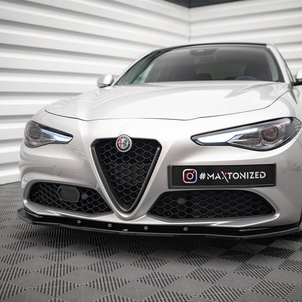 A silver car with sleek headlights and geometric grille stationary in a showroom-like setting with textured walls and floor. The text on a plate reads: "@MAXTONIZED".