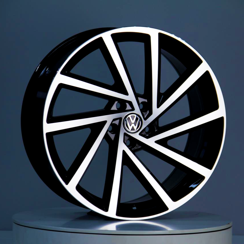 A black and silver alloy wheel, featuring the Volkswagen logo at its center, is displayed upright against a neutral gray background on a reflective surface.