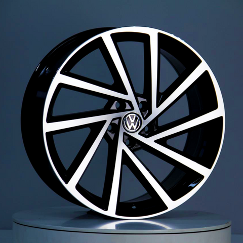 A black and silver alloy wheel, featuring the Volkswagen logo at its center, is displayed upright against a neutral gray background on a reflective surface.