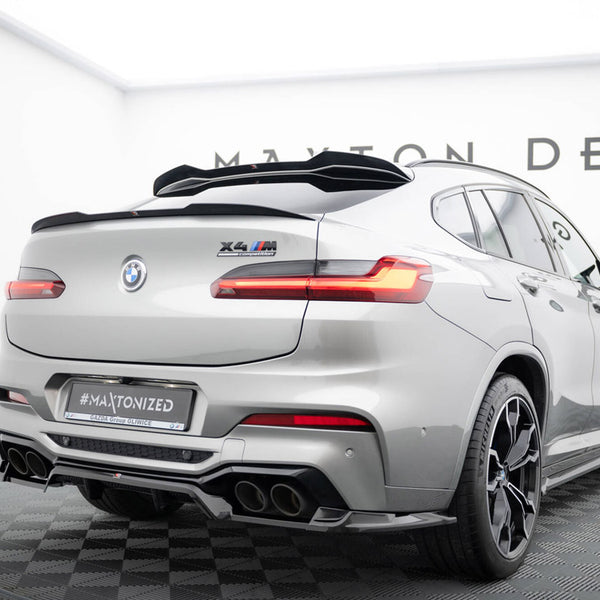 A silver BMW X4 M, featuring a rear spoiler and quad exhausts, is stationary on a checkered platform. Visible text includes "X4 M competition" and "#MAXTONIZED". It's in a showroom with "MAXTON DESIGN" partially visible on the wall.