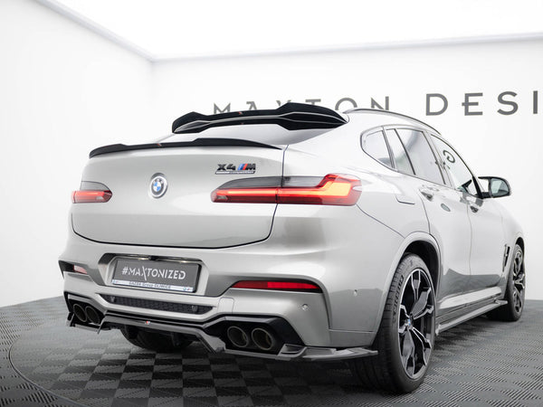 A silver BMW X4 M, featuring a rear spoiler and quad exhausts, is stationary on a checkered platform. Visible text includes "X4 M competition" and "#MAXTONIZED". It's in a showroom with "MAXTON DESIGN" partially visible on the wall.