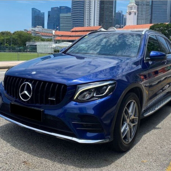 A blue Mercedes-Benz SUV is parked on a city street. Skyscrapers and a clock tower appear in the background, with greenery and a clear sky enhancing the urban setting.