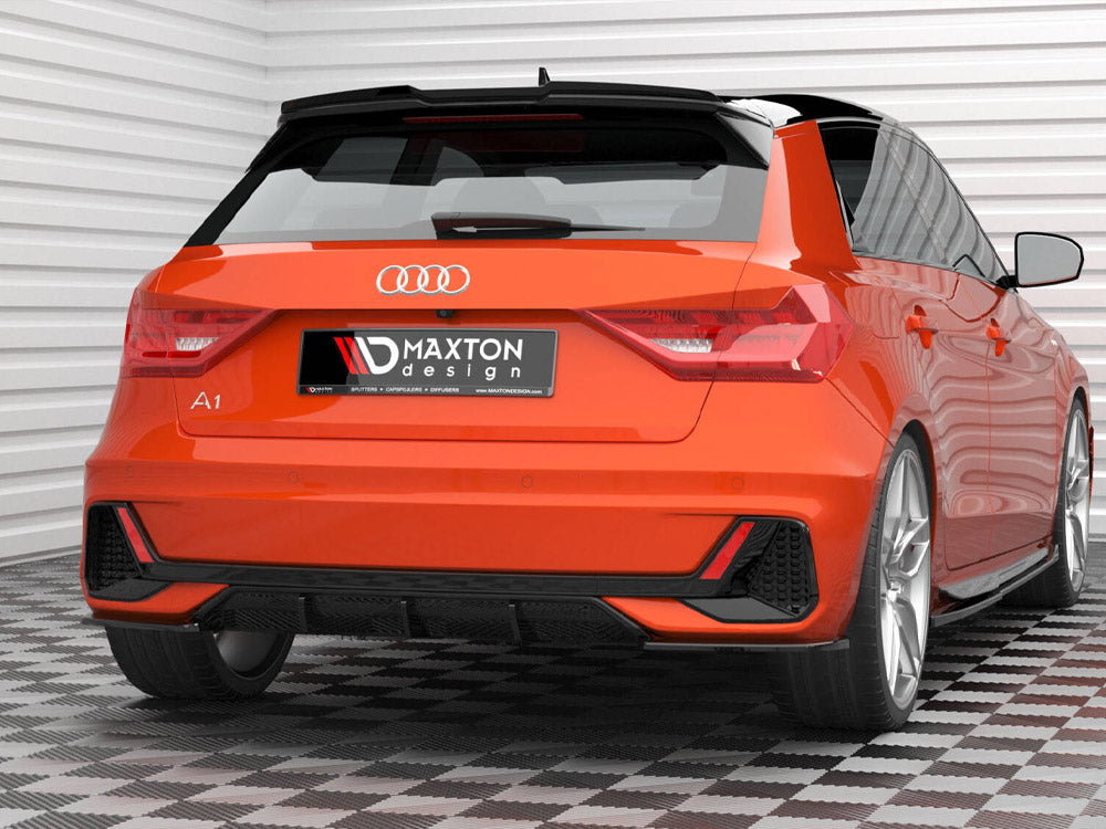 A bright orange Audi A1 is parked in a garage with white paneled walls and checkered flooring. The license plate reads "MAXTON design." The car features sleek, sporty lines.