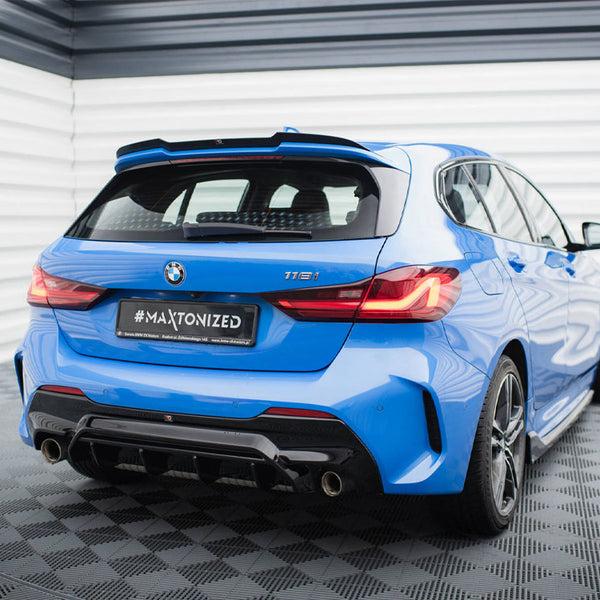 A blue BMW 116i parked, rear view, with illuminated taillights, in a showroom with a geometric-patterned floor and striped walls. License plate reads "#MAXTONIZED".