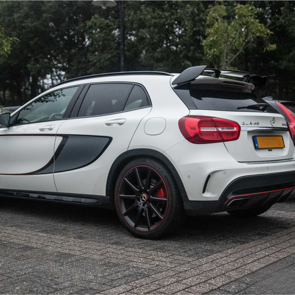 A white car with a sporty design, labeled "GLA 45" and black-and-red accents, is parked on a paved road. Other vehicles and leafy bushes are in the background.