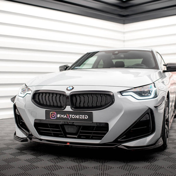 A gray BMW car is parked stationary in a modern showroom with white, geometric walls. The license plate displays Instagram text: "#MAXTONIZED".