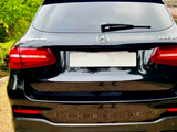 A black Mercedes-Benz GLC 63 AMG parked on a gravel driveway, surrounded by green trees. The rear of the car features four exhaust pipes and red taillights.