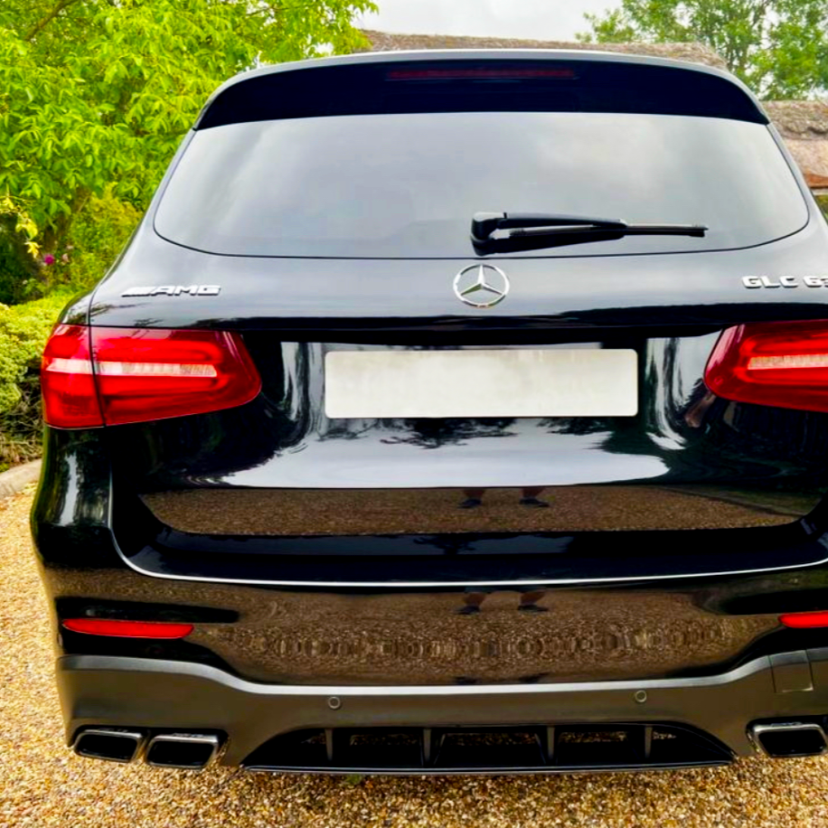A black Mercedes-Benz GLC 63 AMG parked on a gravel driveway, surrounded by green trees. The rear of the car features four exhaust pipes and red taillights.