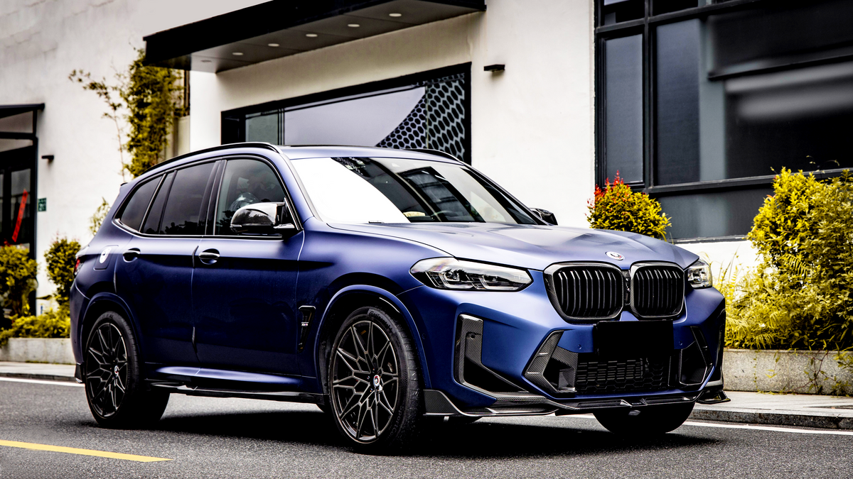 A metallic blue SUV is parked on a street beside a modern building with large windows and greenery, showcasing its sleek design and black wheels.
