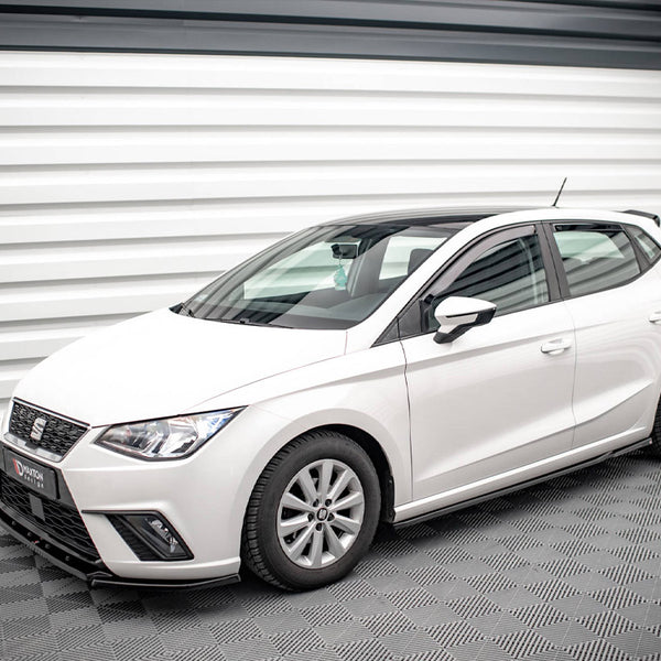 A white hatchback car is parked in a modern, well-lit showroom with patterned flooring and walls.