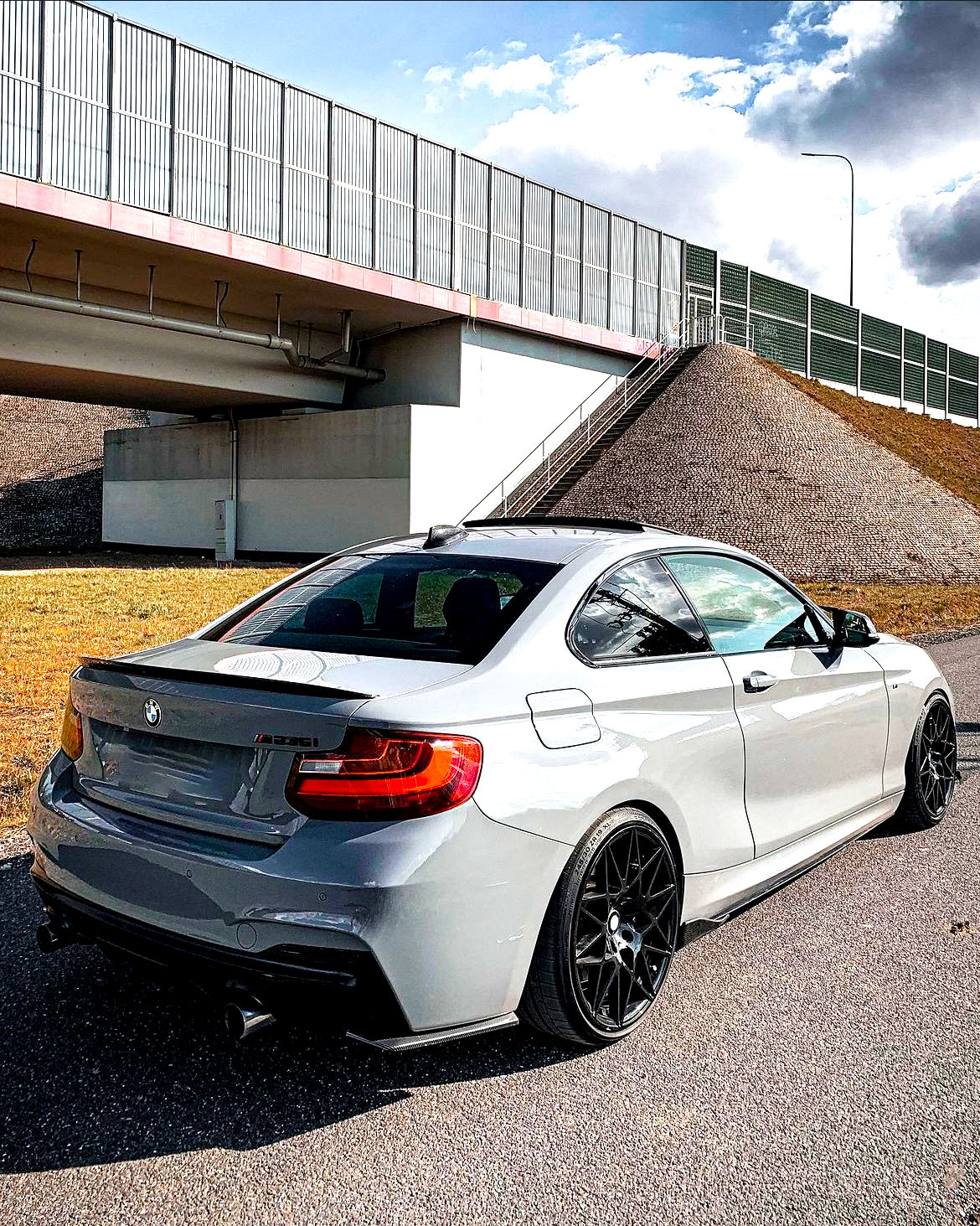 A gray BMW sports car is parked on a road beneath a large bridge with metal railings, near grassy embankments under a partly cloudy sky.