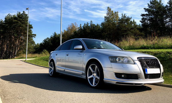 A silver car is parked on a sunlit road beside a grassy area with trees and street lamps in the background. License plate reads "EST".
