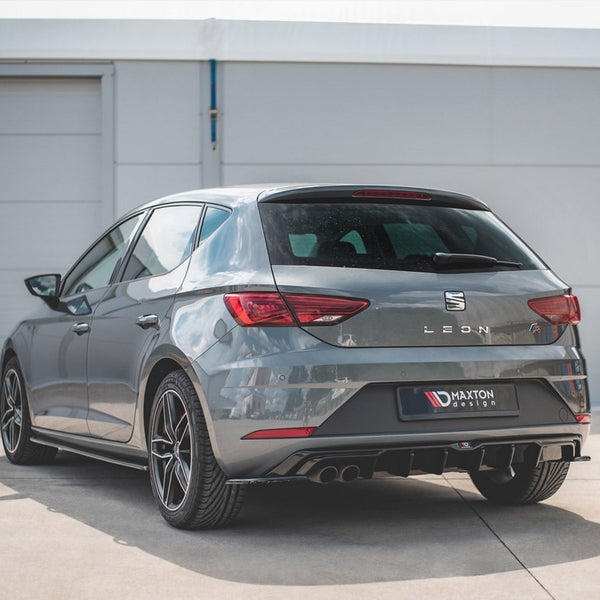 A grey SEAT Leon car is parked on a concrete surface near a white building. The rear features a Maxton Design diffuser and visible dual exhausts.