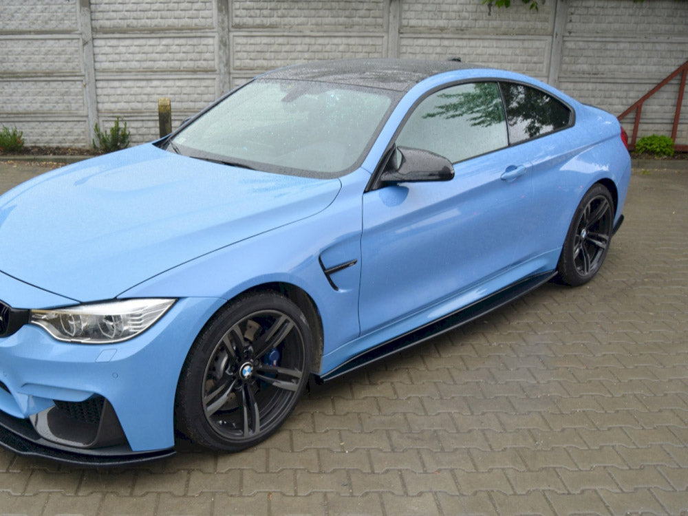A blue sports car is parked on a wet, paved area, with a gray brick wall and some greenery in the background.
