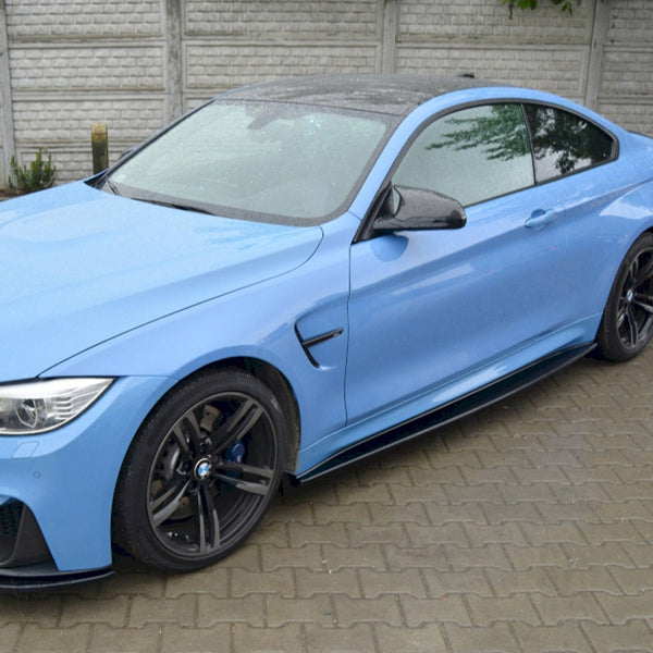 A blue sports car is parked on a wet, paved area, with a gray brick wall and some greenery in the background.