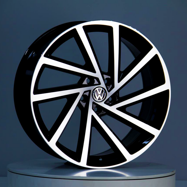 A black and white alloy wheel, featuring a Volkswagen logo in the center, stands on a round podium against a gray background.