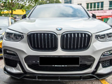 A white BMW car is parked, displaying its distinctive grille and headlights. It's situated in an outdoor parking area, with other cars and trees visible in the background.
