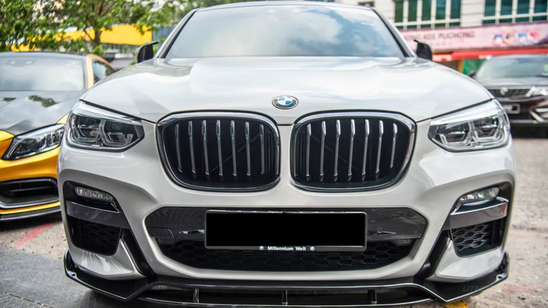 A white BMW car is parked, displaying its distinctive grille and headlights. It's situated in an outdoor parking area, with other cars and trees visible in the background.