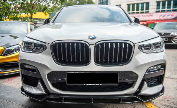 A white BMW car is parked, displaying its distinctive grille and headlights. It's situated in an outdoor parking area, with other cars and trees visible in the background.