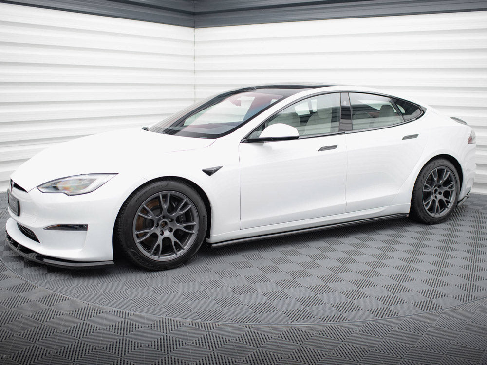 A white electric car is parked on a textured, dark floor within a bright, windowless room.