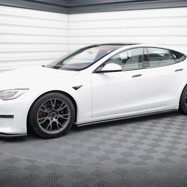 A white electric car is parked on a textured, dark floor within a bright, windowless room.