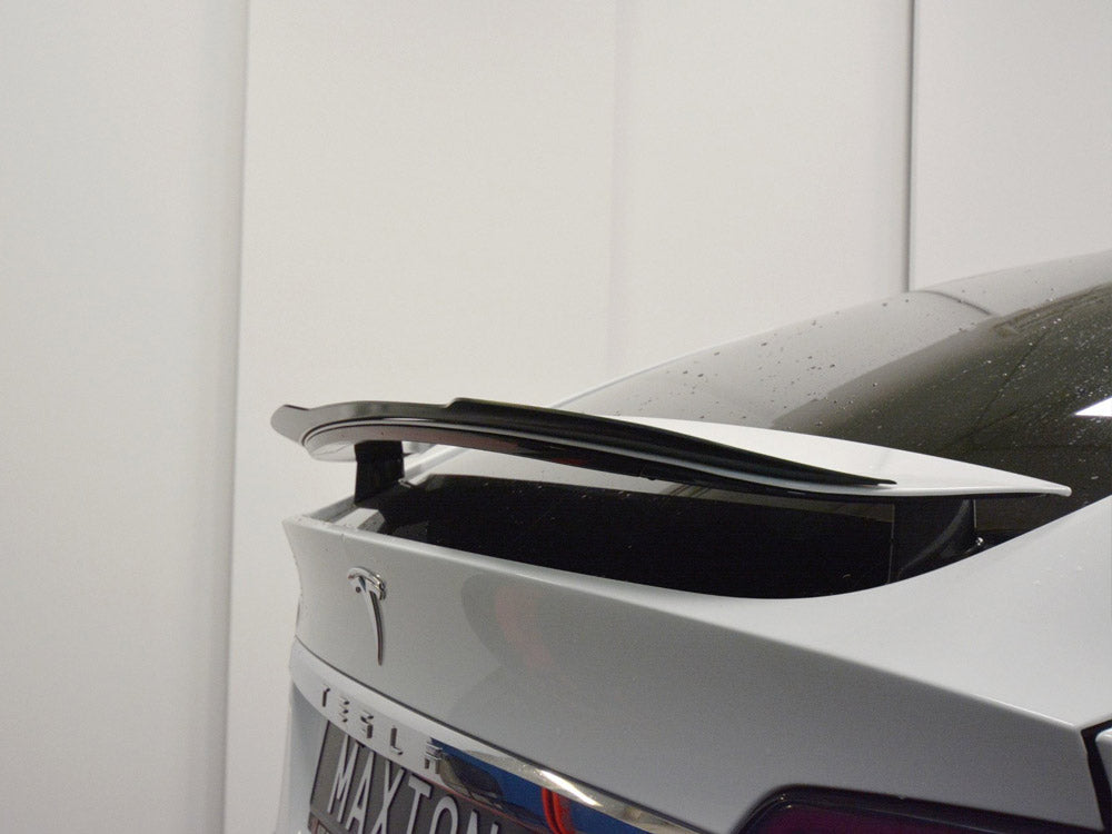 A car's rear end features a black spoiler on a white polished surface, with "MA-XTON" visible on the license plate. The background is a plain white wall.
