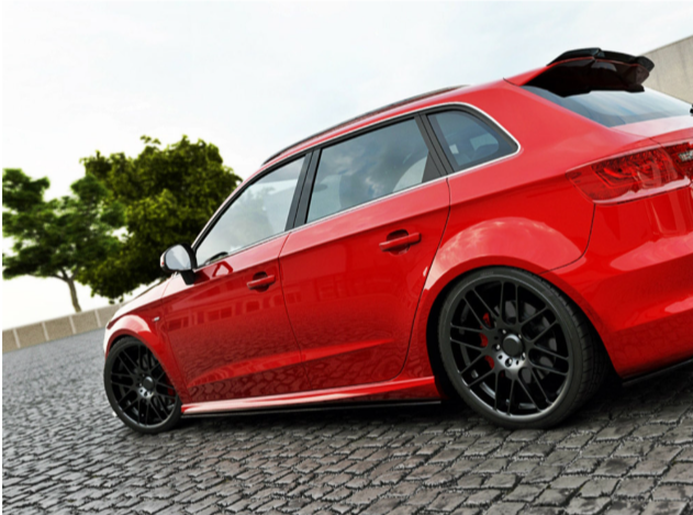 A red car is parked on a cobblestone surface. It has black alloy wheels and a rear spoiler. In the background, there are trees and a low concrete wall.