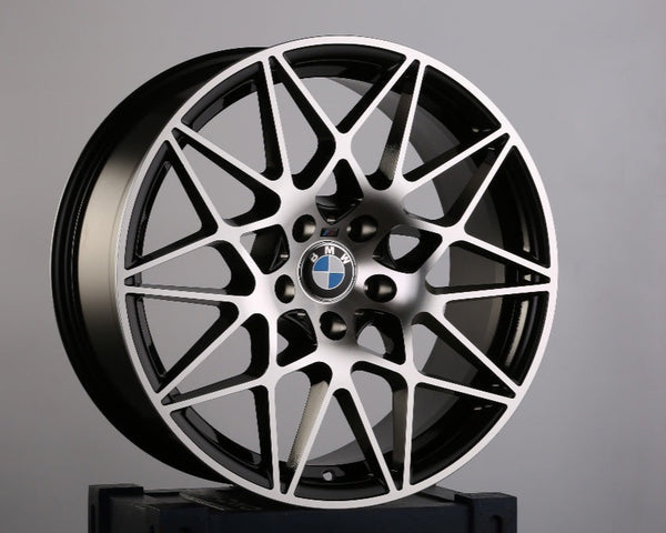 A BMW car rim with an intricate, geometric design stands upright on a dark blue box emblazoned with the letters "GA" and a partial Union Jack in a plain gray environment.