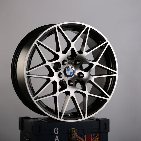 A BMW car rim with an intricate, geometric design stands upright on a dark blue box emblazoned with the letters "GA" and a partial Union Jack in a plain gray environment.