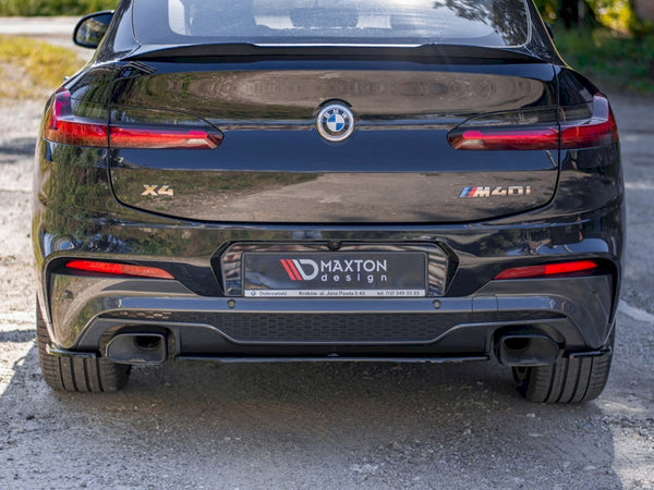 A black BMW X4 M40i is parked on a gravel surface. The license plate reads "MAXTON design." The car's rear includes dual exhausts and prominent taillights.