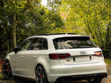 A white Audi car is parked, showcasing sleek black-rimmed wheels, amidst lush green trees and scattered fallen leaves, creating a serene, natural setting. The rear logo reads 
