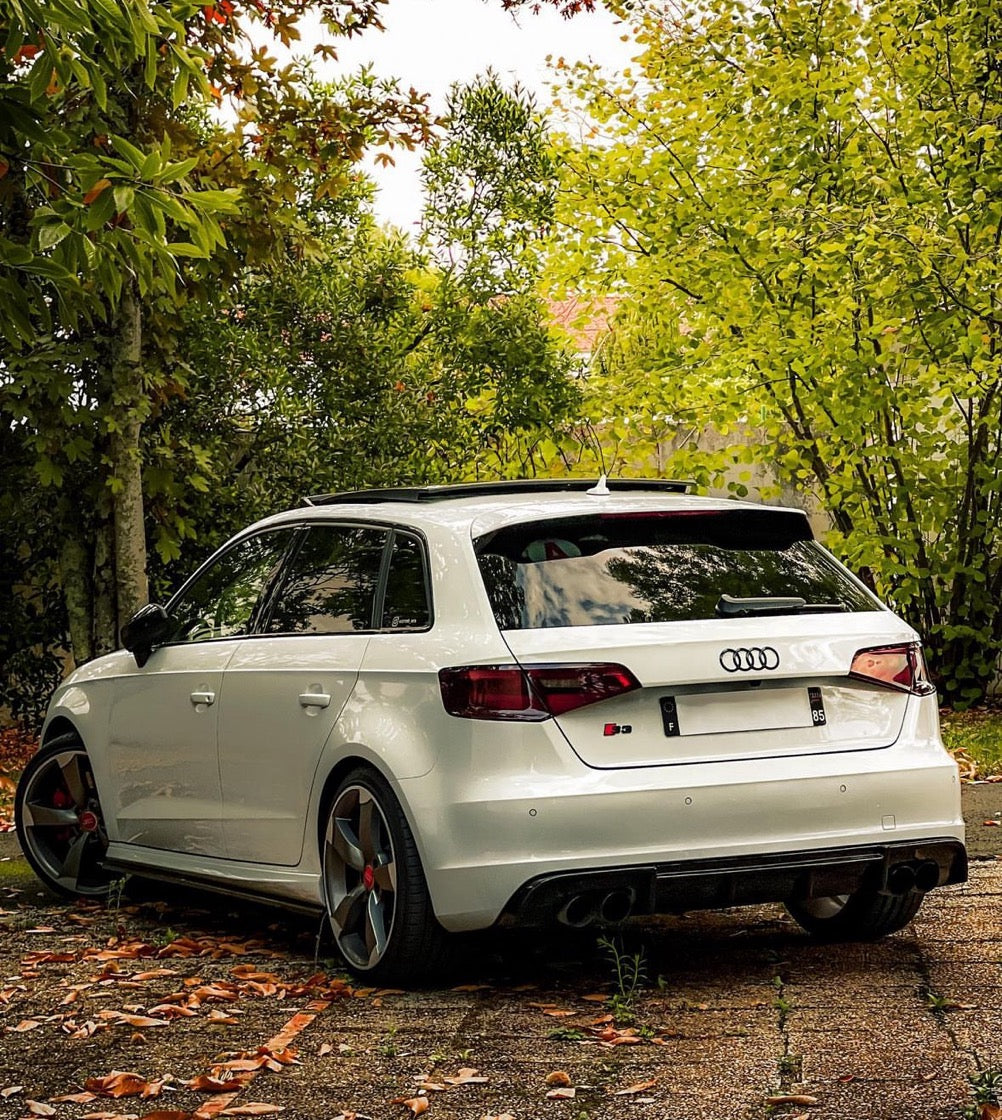 A white Audi car is parked, showcasing sleek black-rimmed wheels, amidst lush green trees and scattered fallen leaves, creating a serene, natural setting. The rear logo reads "S3."
