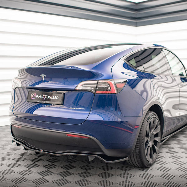 A blue Tesla Model Y, displaying a "DUAL MOTOR" badge, is parked in a well-lit, modern garage with checkered flooring. The license plate reads: "@#MAATONIZED YELRAX.COM".