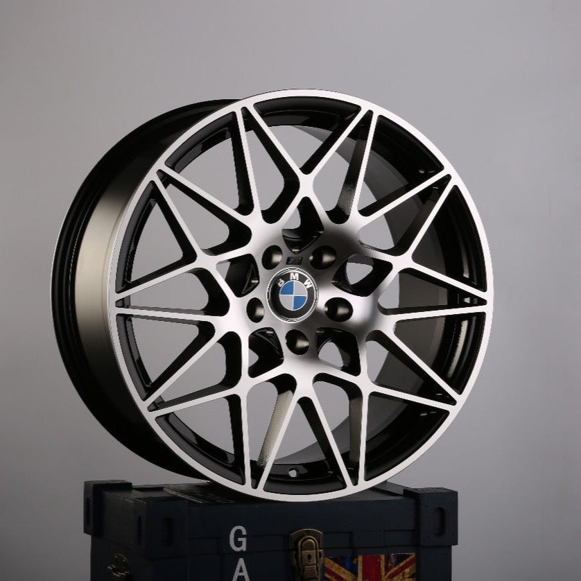 A BMW wheel with intricate multi-spoke design rests atop a dark-blue crate, featuring a faint Union Jack pattern and partial text "GA" visible, against a plain grey background.