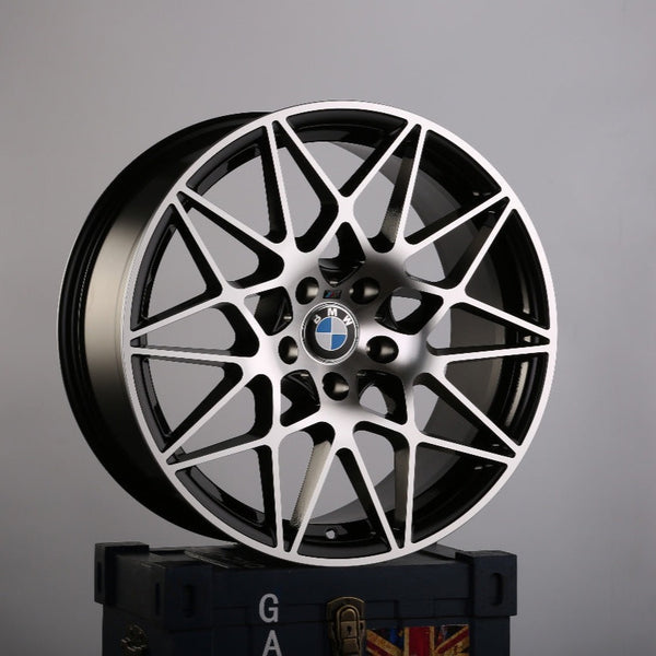 A BMW wheel with intricate multi-spoke design rests atop a dark-blue crate, featuring a faint Union Jack pattern and partial text "GA" visible, against a plain grey background.