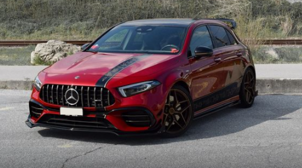 A red Mercedes-Benz car is parked on a paved road, featuring black stripes, a prominent grille, and dark alloy wheels. Behind are grass and train tracks, creating an urban backdrop.