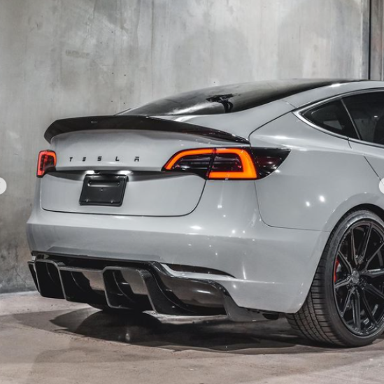 A gray Tesla car is parked indoors, featuring illuminated tail lights and a sleek design. The letters "T E S L A" are visible on the rear, against a concrete wall.
