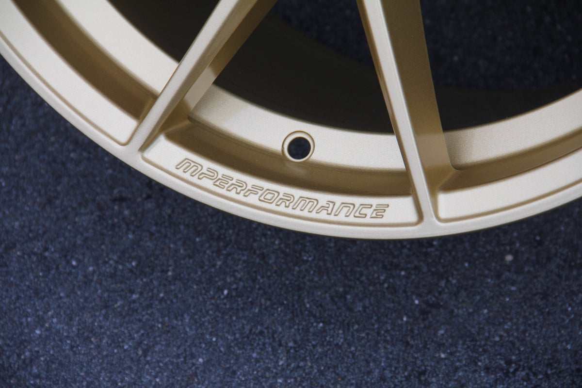 A gold car rim with the text "MPERFORMANCE" engraved, shown close-up on a textured black surface.