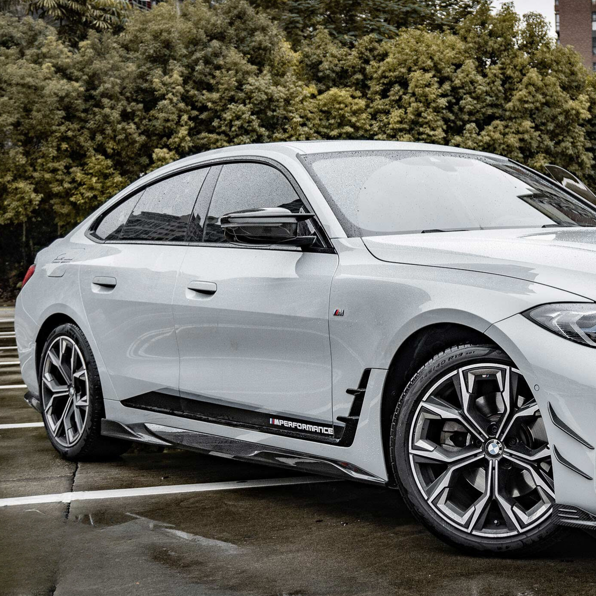 A silver car with sleek, modern design parked in an outdoor setting; trees form the background. The side reads "M Performance," emphasizing high performance. Light rain leaves droplets on the surface.