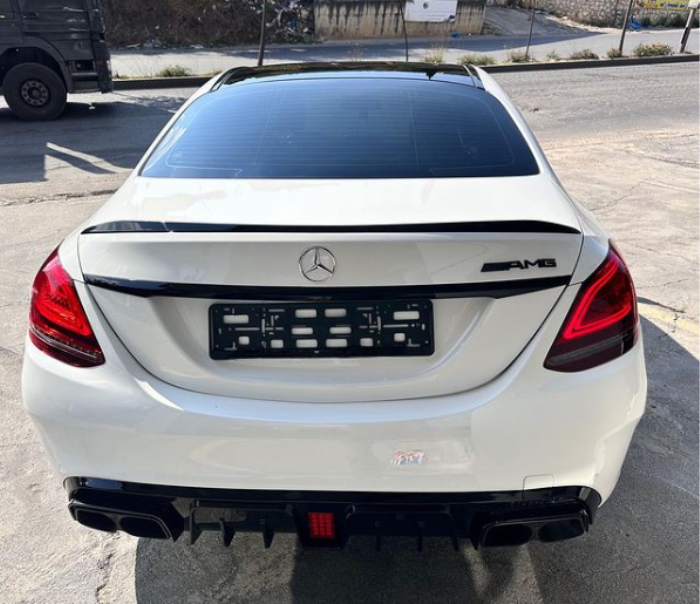 A white Mercedes AMG car is parked on a concrete surface, viewed from the rear. The context includes an empty license plate holder, red taillights, and a nearby road with greenery.