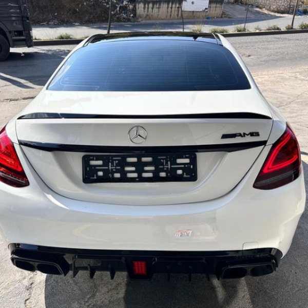 A white Mercedes AMG car is parked on a concrete surface, viewed from the rear. The context includes an empty license plate holder, red taillights, and a nearby road with greenery.