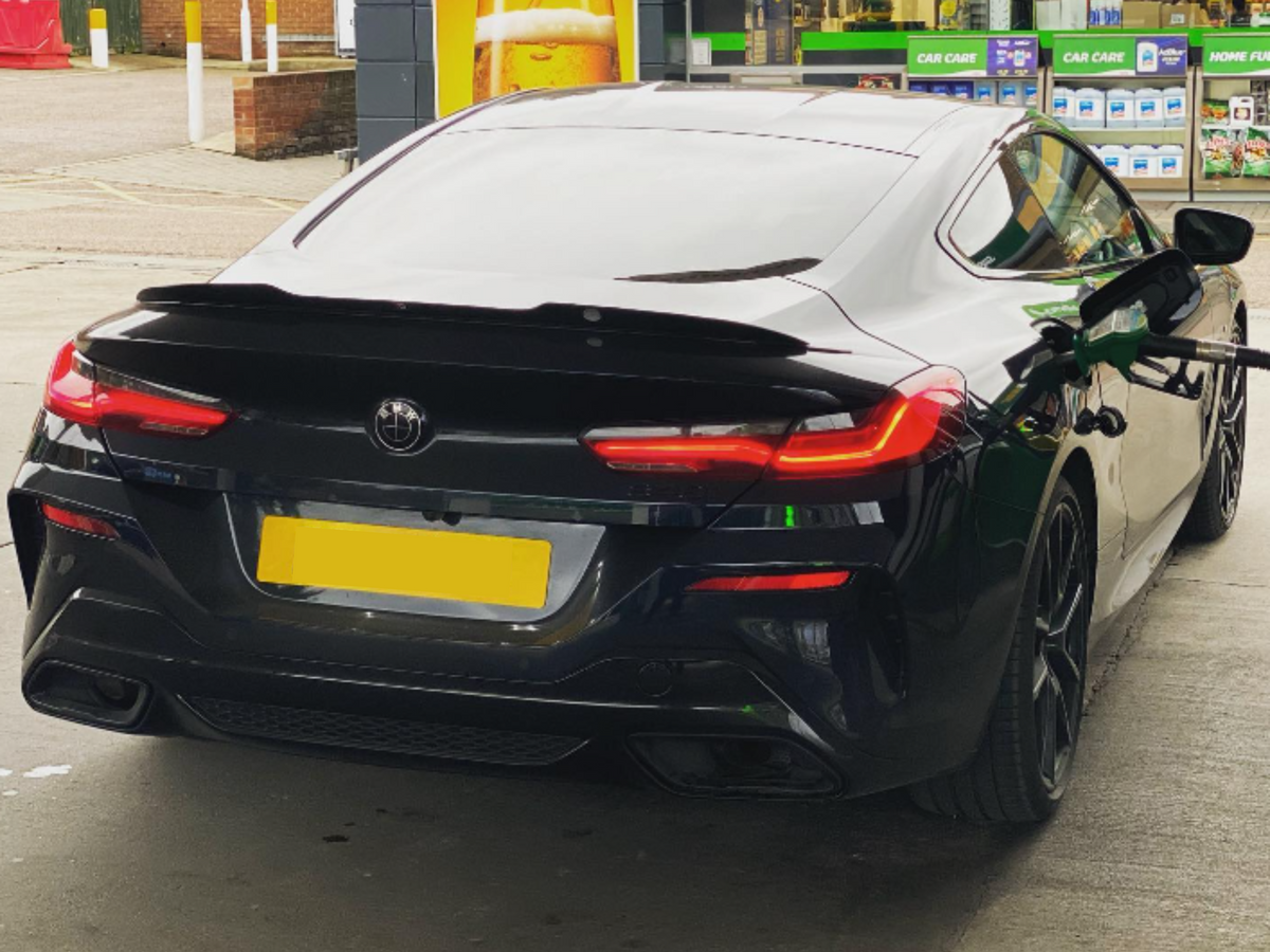 A black sports car is refueling at a gas station. The car's rear lights are illuminated, and shelves with automotive products are visible in the background.