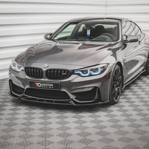 A sleek, gray BMW sports car with illuminated headlights sits on a checkered floor in a bright, white-paneled room. A license plate reads: "MAXTON design."