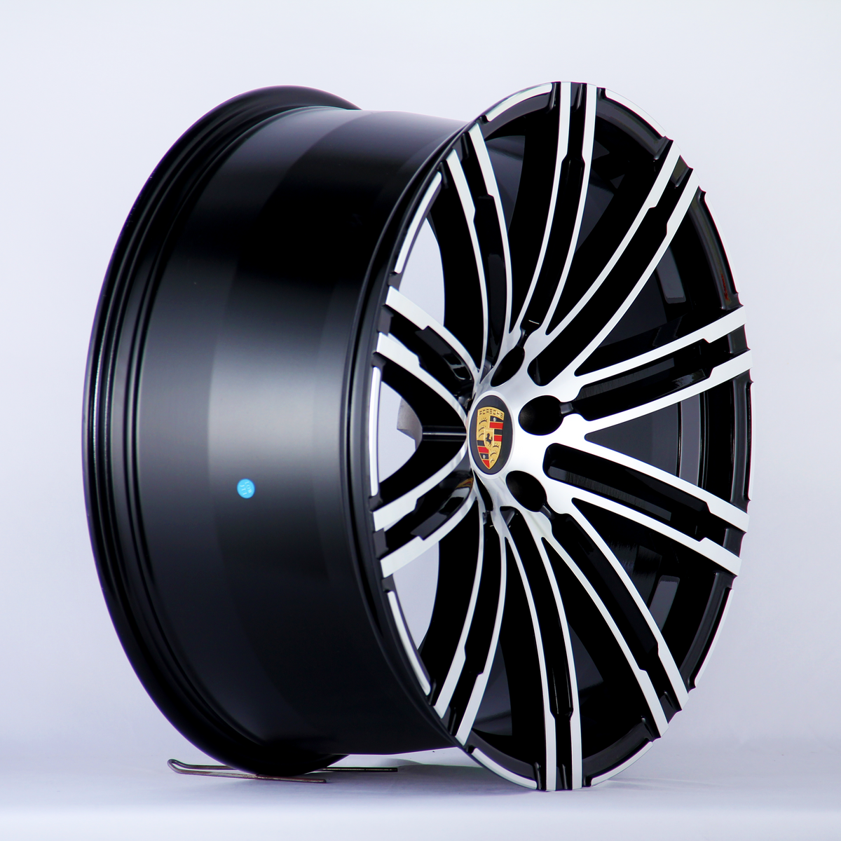 A glossy black and silver alloy wheel is angled to show its intricate spoke design, featuring a central emblem with the Porsche logo. The background is plain white.