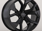 A black, glossy alloy wheel with a dynamic, angular spoke design. The Audi logo is at its center. The wheel is set against a plain, light gray background.