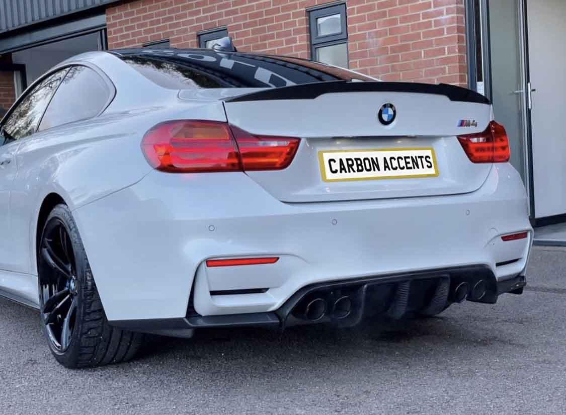 A white BMW car is parked, displaying its black wheels and dual exhausts. It has a license plate reading "CARBON ACCENTS." The setting features a brick building and a glass door.