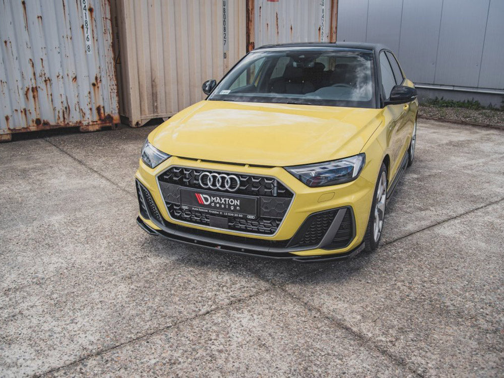 A yellow car is parked on a concrete surface near rusted shipping containers. The front license plate reads "MAXTON design."